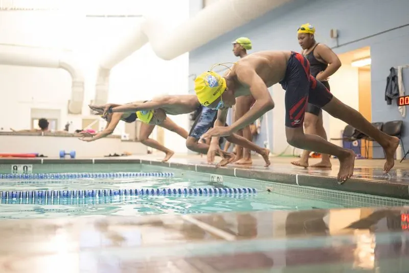 سباحون شباب يرتدون قبعات سباحة صفراء يقفزون نحو الماء في مسبح داخلي، يعكس روح المنافسة والانتماء لفريق ماكوس للسباحة.