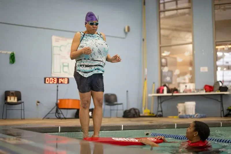 امرأة ترتدي نظارات سباحة وقبعة، تستعد للقفز إلى الماء بينما تراقبها سباحة أخرى في مسبح، تعكس روح التعلم والتمكين في السباحة.