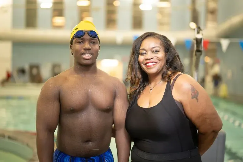 شاب يرتدي قبعة سباحة صفراء ونظارات سباحة يقف بجانب والدته في مركز السباحة. خلفهما، يظهر حوض السباحة وأعلام ملونة.