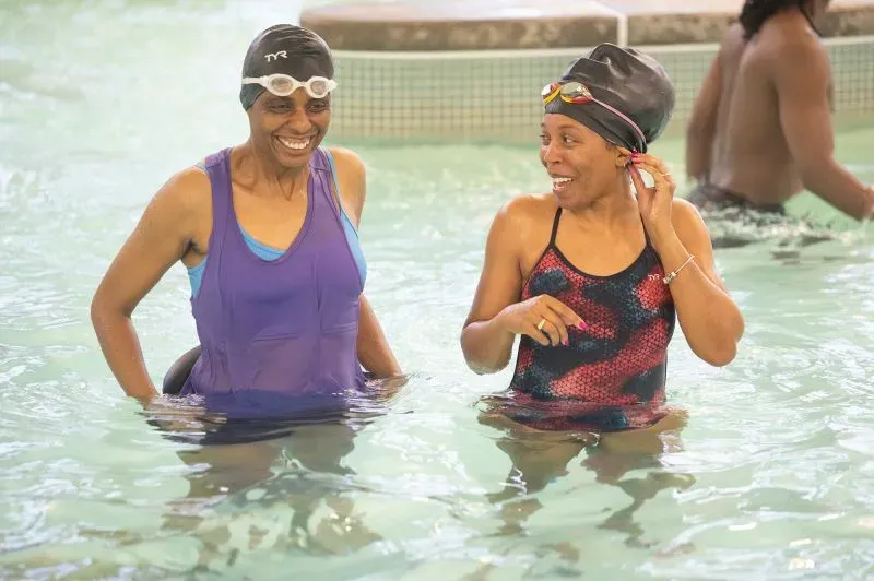 امرأتان تتبادلان الضحك في مسبح، ترتديان ملابس سباحة وقبعات سباحة، بينما يسبح آخرون في الخلفية، مما يعكس أهمية السباحة في المجتمع.