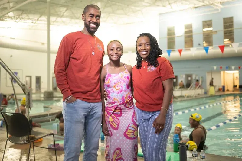 عائلة تدعم السباحة، مع شاب في منتصف الصورة، في مركز ترفيهي. يظهر خلفهم حمام سباحة ومشاركون يرتدون قبعات سباحة صفراء.