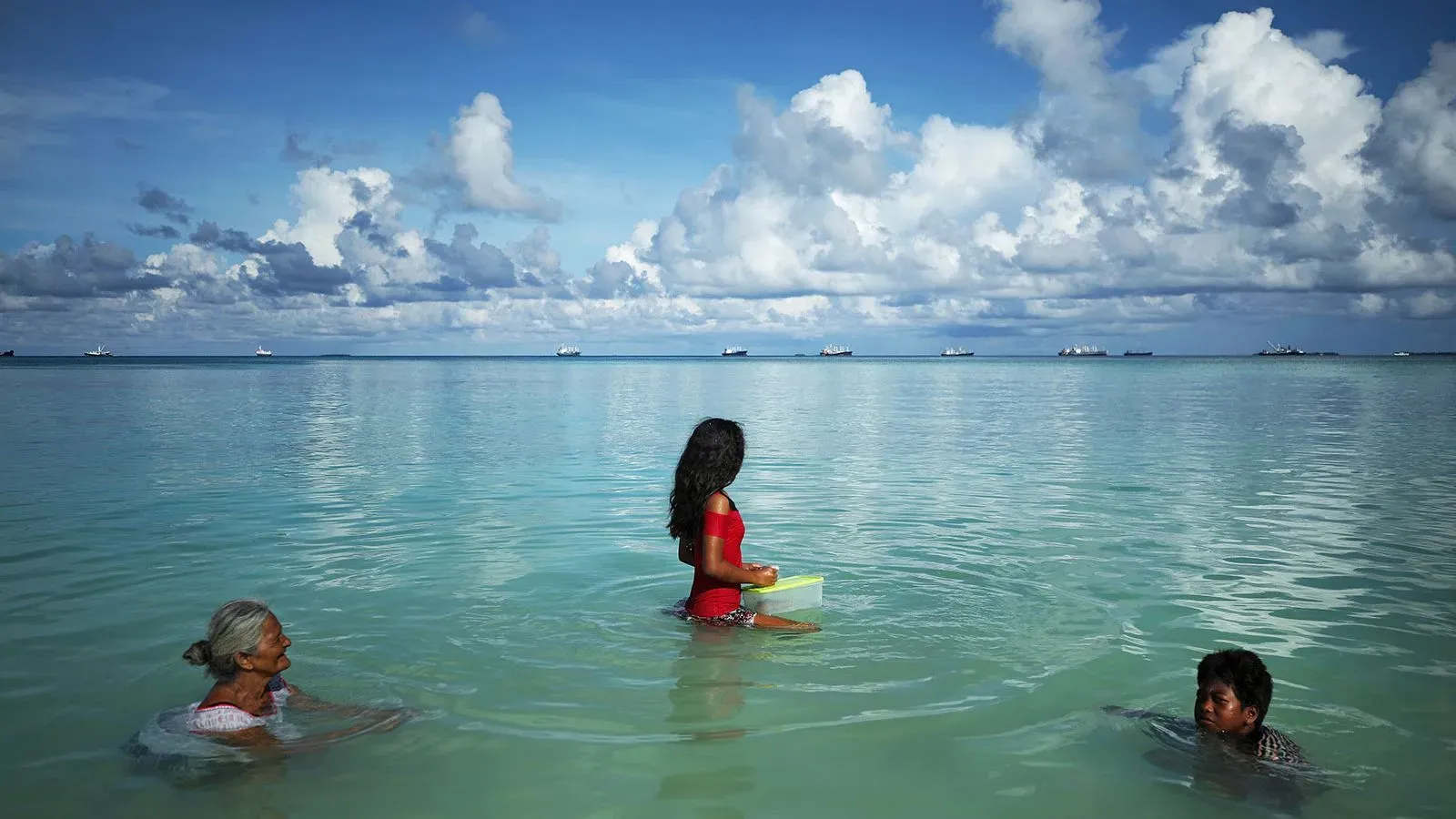 امرأتان وفتاة صغيرة في مياه بحر توفالو الضحلة، مع سفن تبحر في الخلفية، تعكس تأثير ارتفاع منسوب المياه على المجتمع المحلي.