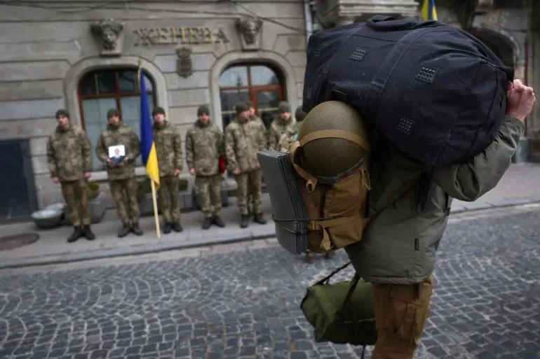 جندي أوكراني يحمل حقيبة كبيرة ويرتدي خوذة، بينما يقف جنود آخرون في صف خلفه، في مشهد يعكس الوضع العسكري في أوكرانيا.