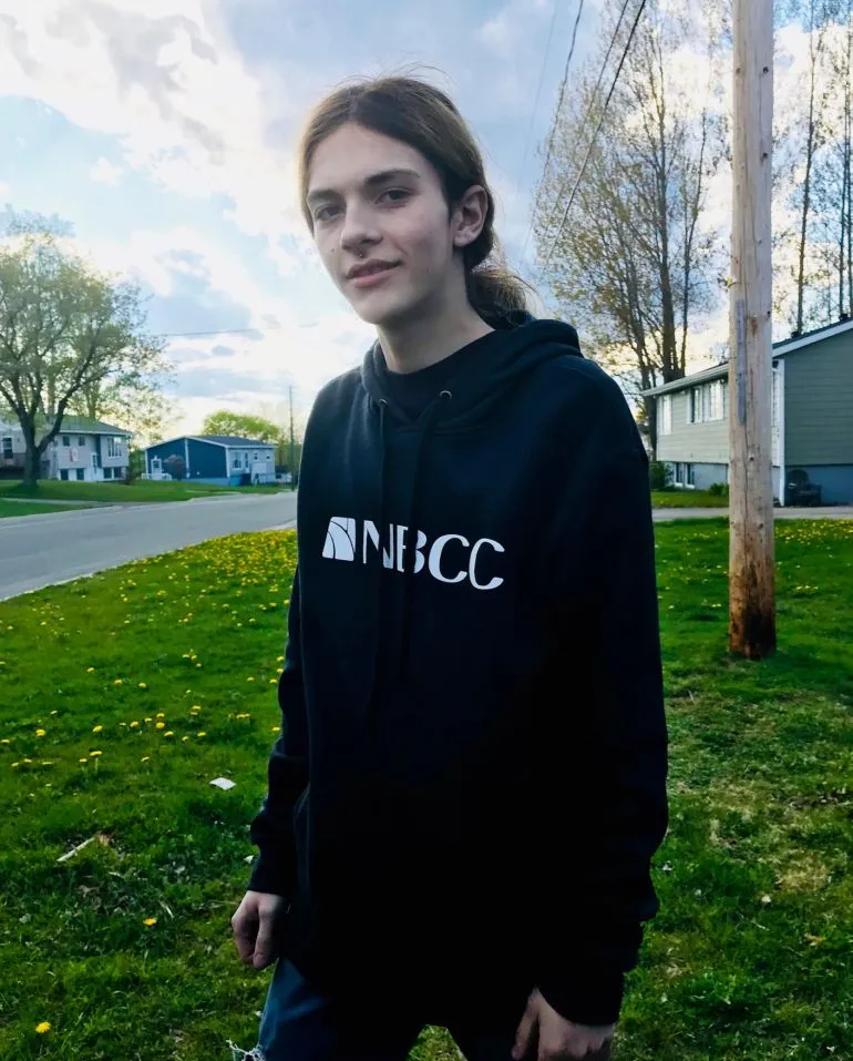 صورة لشابة ترتدي سترة سوداء تحمل شعار NBCC، تقف في حديقة مع أشجار وخلفية سماء زرقاء، تعكس لحظة من الهدوء قبل مأساة.