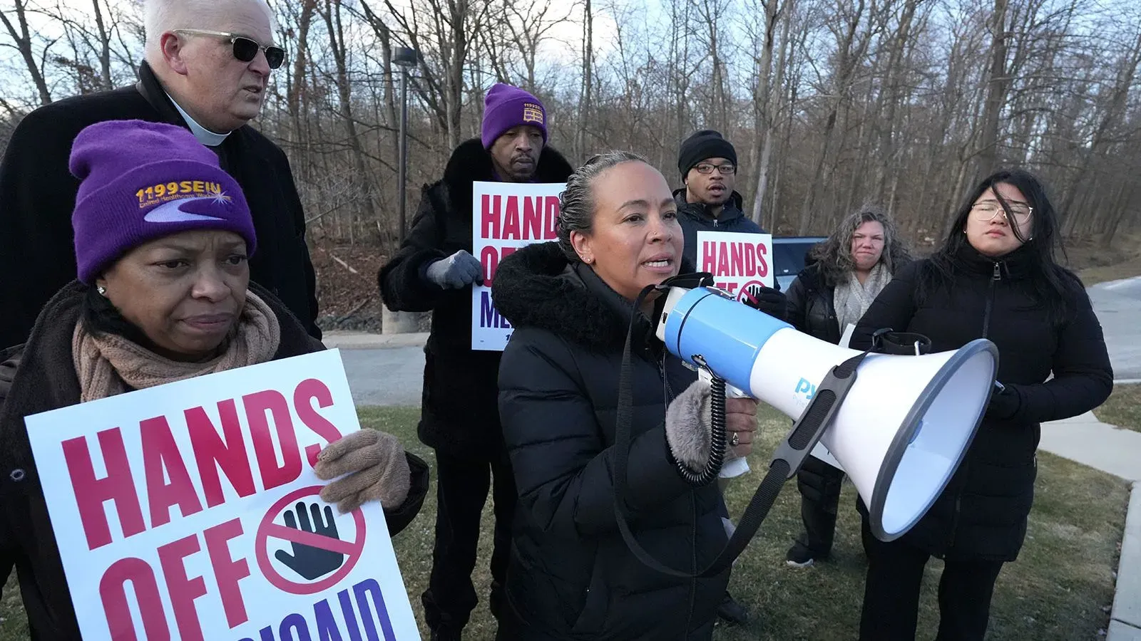 محتجون يحملون لافتات تطالب بحماية برنامج Medicaid، مع امرأة تتحدث عبر ميكروفون، في إطار جهود الدفاع عن الرعاية الصحية.