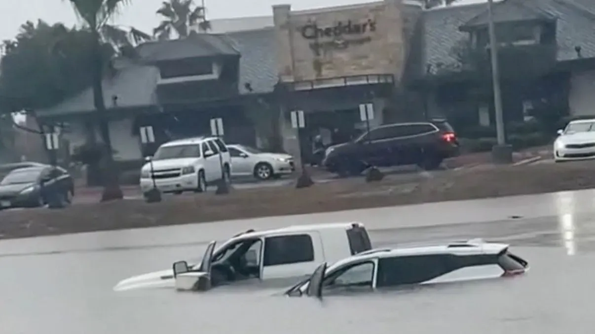 فيضانات شديدة تغمر سيارات في منطقة تجارية بتكساس، مع وجود مطعم "تشيدارز" في الخلفية، نتيجة للعواصف الرعدية.