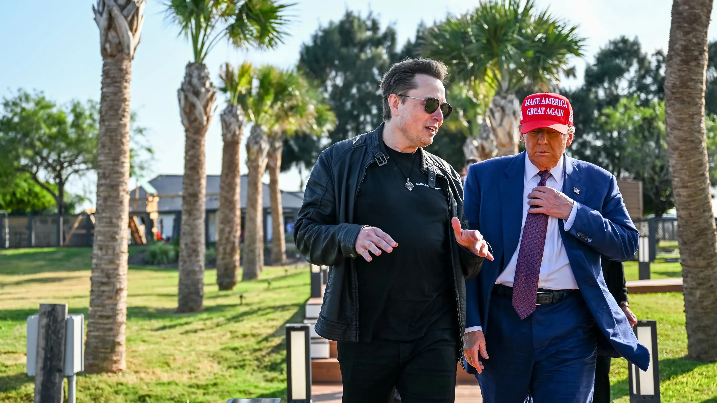 إيلون ماسك ودونالد ترامب يتحدثان في حديقة، مع أشجار النخيل في الخلفية، حيث يتناولان قضايا الحكومة الأمريكية.