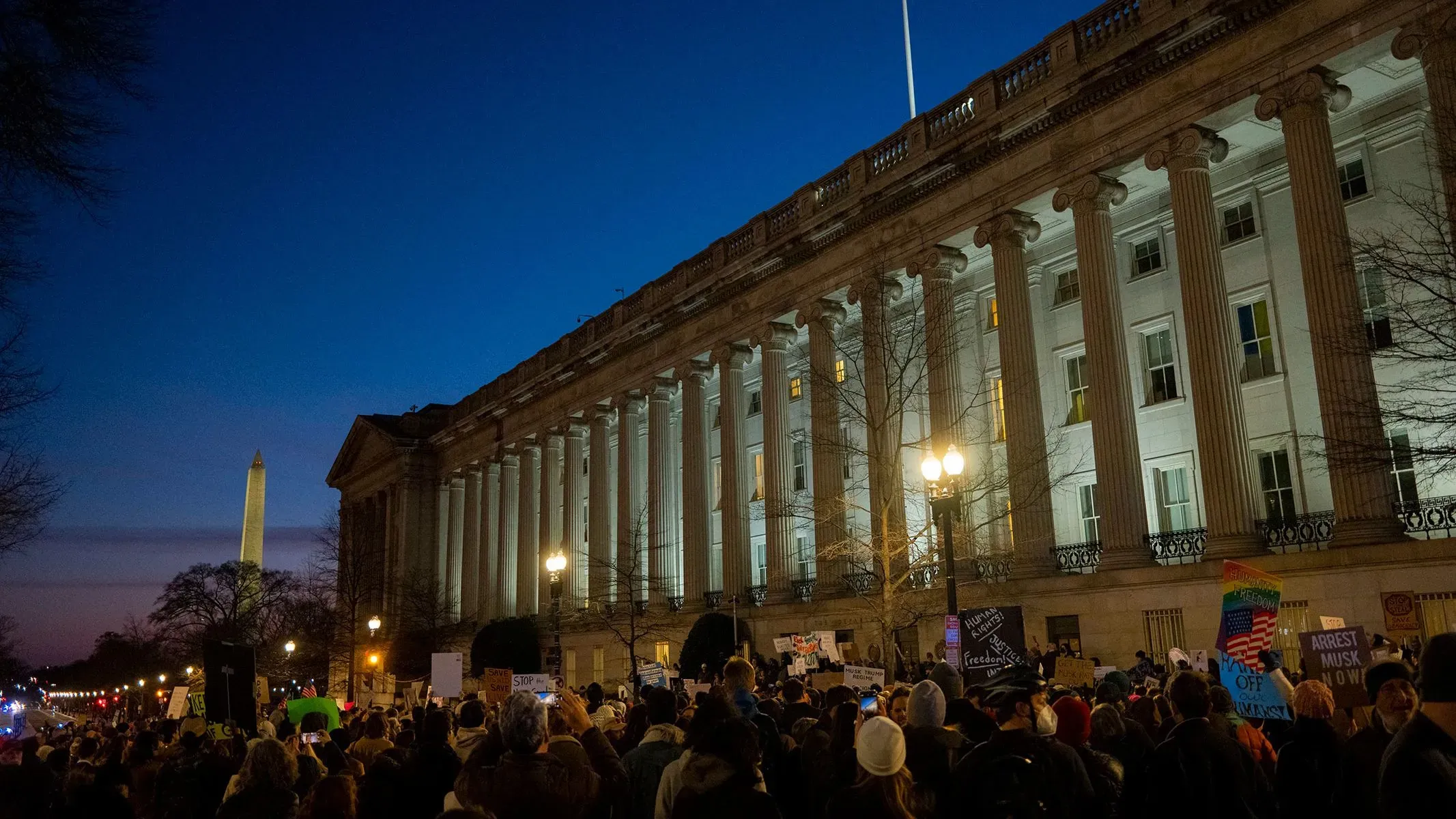 تجمع حشود من المتظاهرين أمام مبنى وزارة الخزانة الأمريكية، مع وجود العلم الأمريكي وواشنطن ميموريال في الخلفية، خلال غروب الشمس.