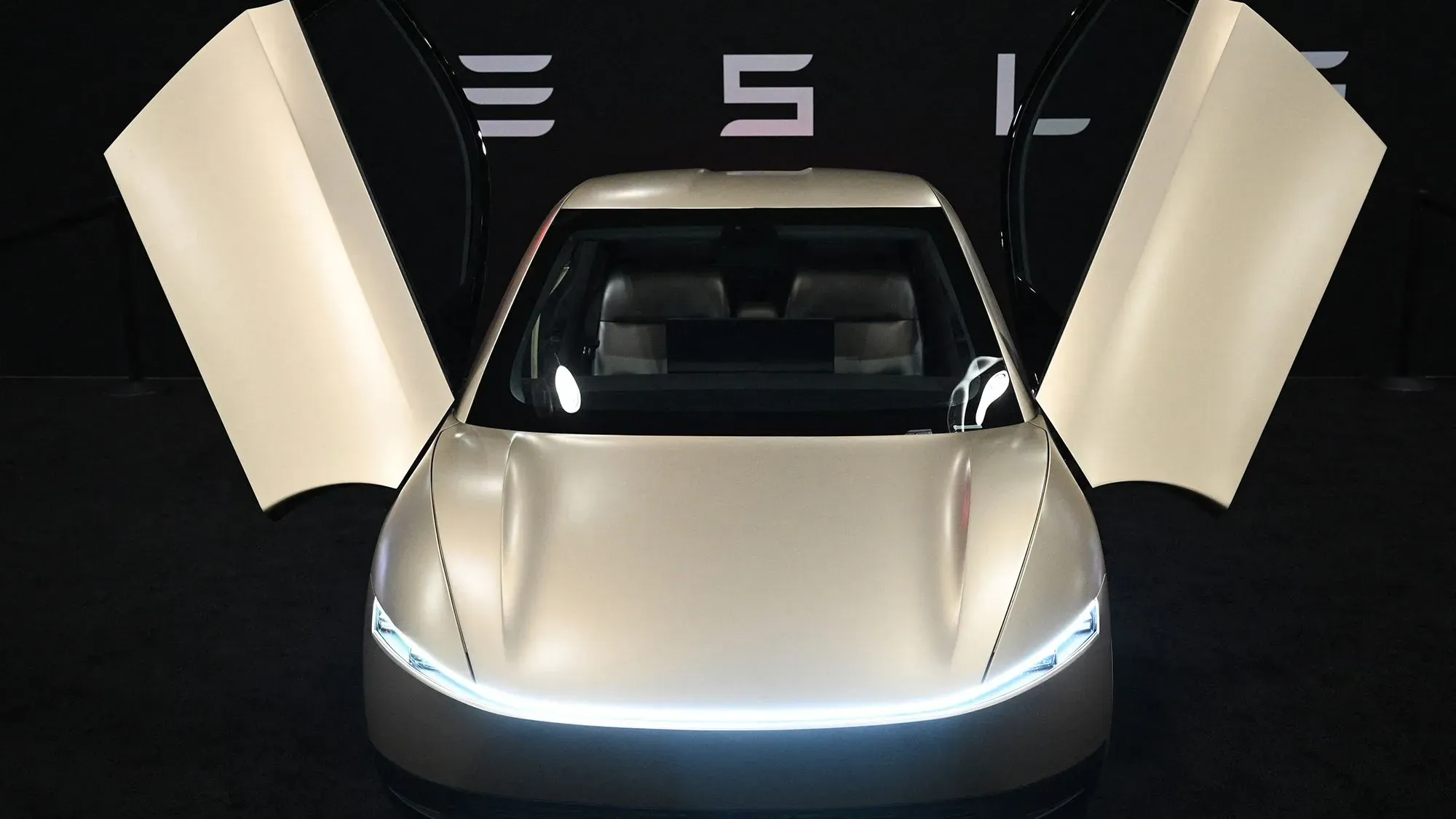 سيارة تسلا الكهربائية مع أبواب تفتح لأعلى، تعرض تصميمها العصري في حدث ترويجي، مع خلفية تحمل شعار تسلا.