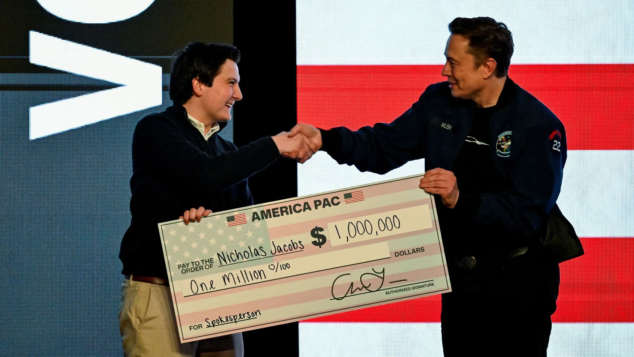 إيلون ماسك يتسلم شيكًا بقيمة مليون دولار من شخص آخر في حدث انتخابي، وسط خلفية تحمل الألوان الوطنية الأمريكية.