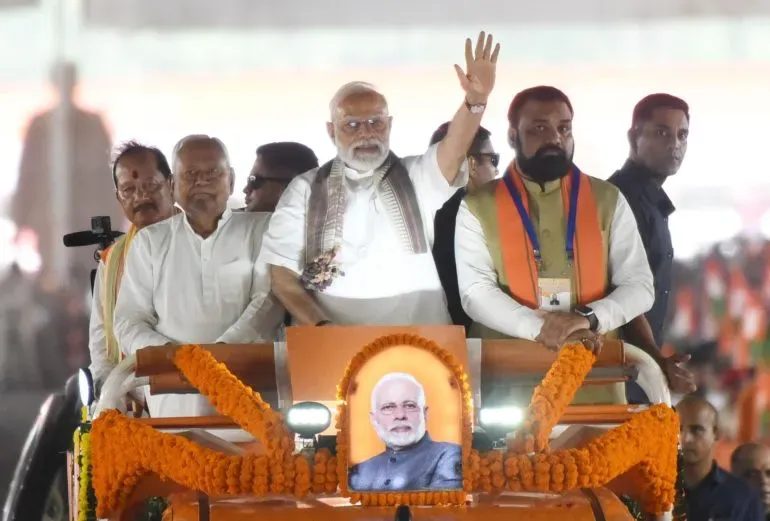 رئيس الوزراء الهندي ناريندرا مودي يلوح بيده خلال تجمع انتخابي في كيشانغانج، مع صورة له على المنصة، وسط حشود من المؤيدين.