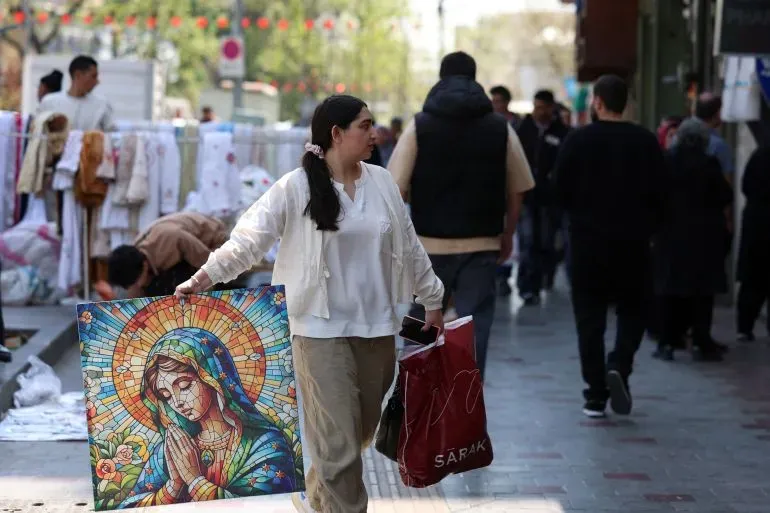 امرأة تحمل لوحة زجاجية ملونة تصور السيدة العذراء، تسير في شارع مزدحم في طهران، بينما يتجول الناس في الخلفية.