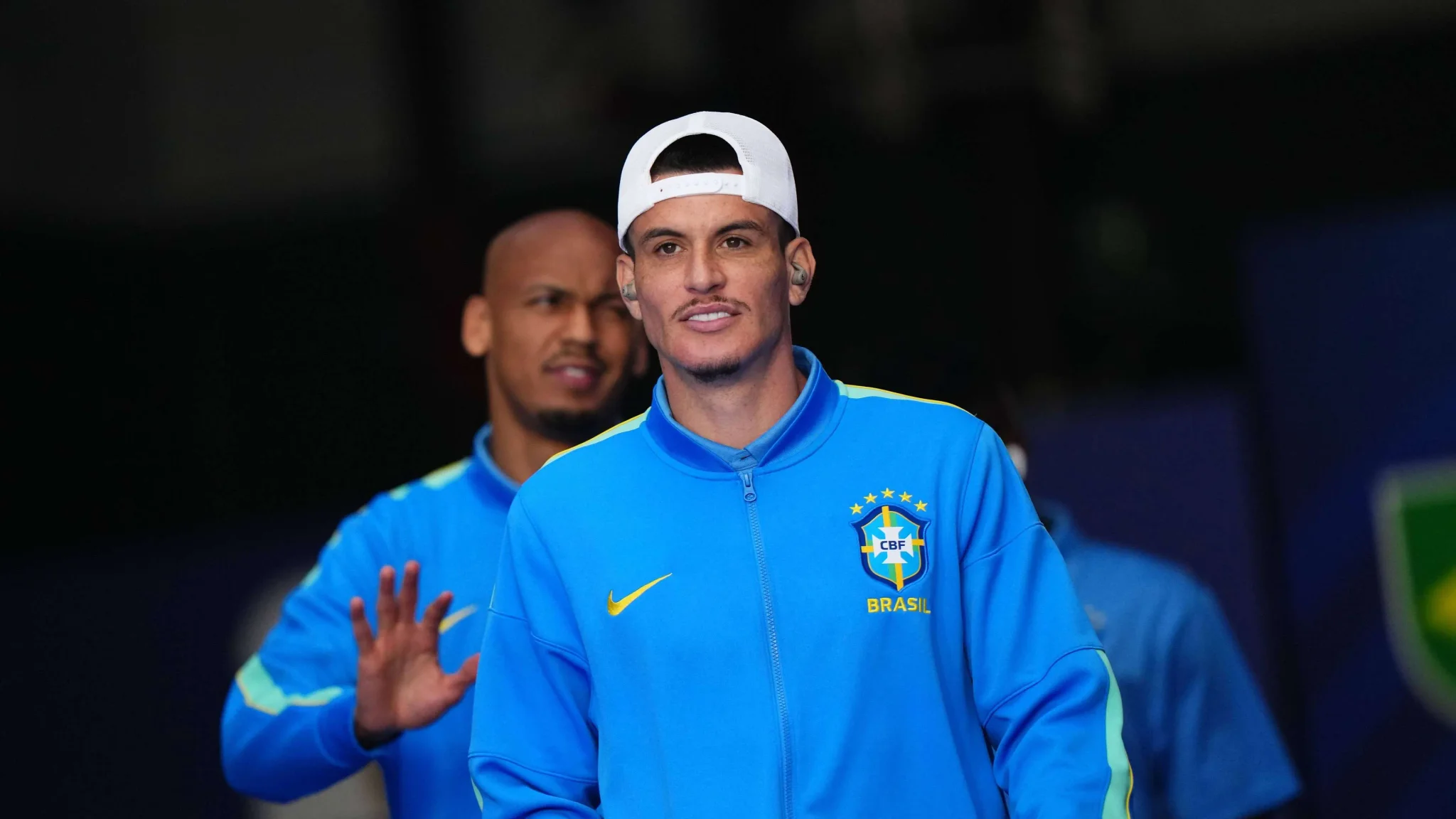 المدافع البرازيلي روجر إيبانيز يرتدي زي منتخب البرازيل الأزرق مع قبعة بيضاء، مع خلفية مظللة ولاعبين آخرين.