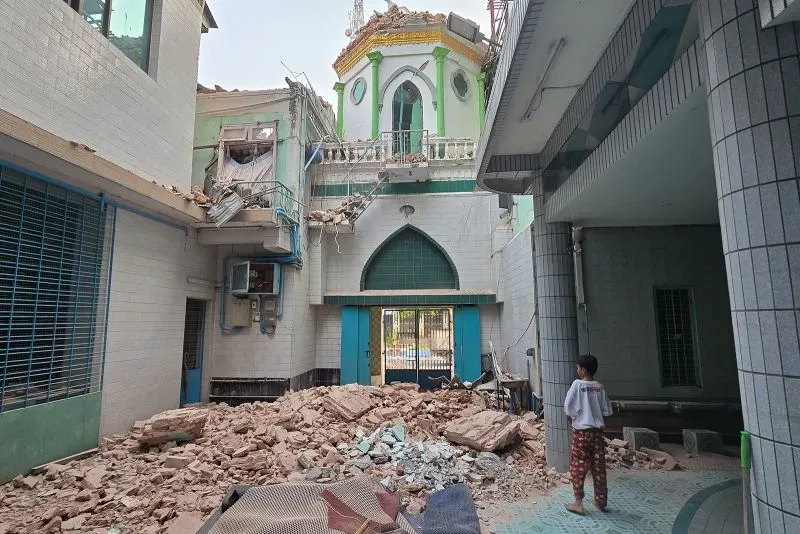 تظهر الصورة آثار الزلزال المدمر في ميانمار، مع أنقاض مبنى ومشاهد دمار واضح، بينما يقف طفل يتأمل الوضع.