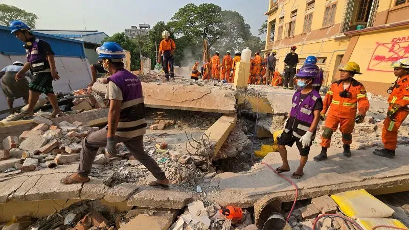 فرق إنقاذ تعمل في موقع الزلزال بميانمار، مع وجود أنقاض مبانٍ مدمرة في الخلفية، تعكس حجم الكارثة الإنسانية.