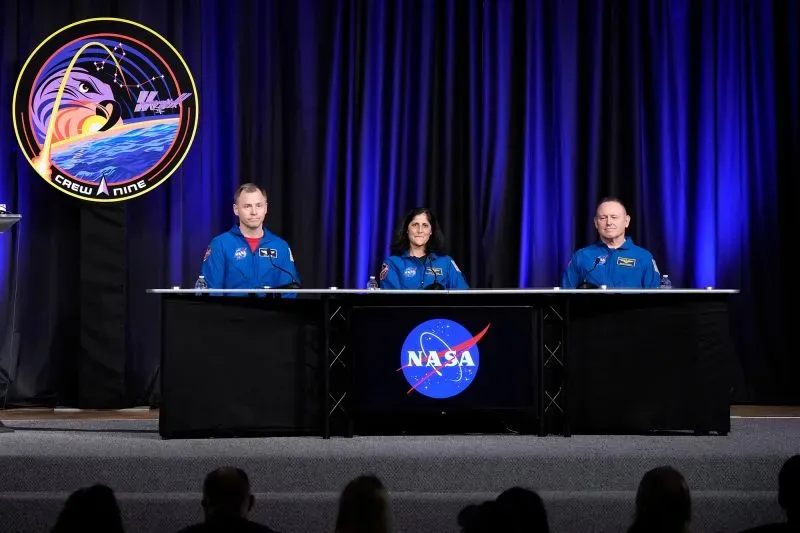 مؤتمر صحفي لرواد الفضاء ويليامز، ويلمور، وهيغ، يتحدثون عن تجربتهم في مهمة كرو-9 مع شعار ناسا خلفهم.