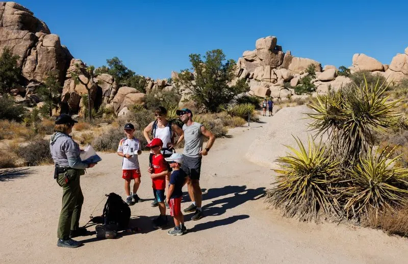 مجموعة من الزوار، بينهم أطفال، يتلقون إرشادات من مرشد في متنزه وطني، مع مناظر طبيعية صخرية في الخلفية.