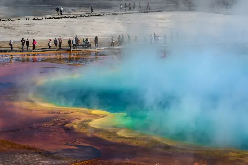 مجموعات من الزوار يستمتعون بمشهد نافورة المياه الساخنة الملونة في متنزه يلوستون الوطني، مع بخار يتصاعد من المياه.