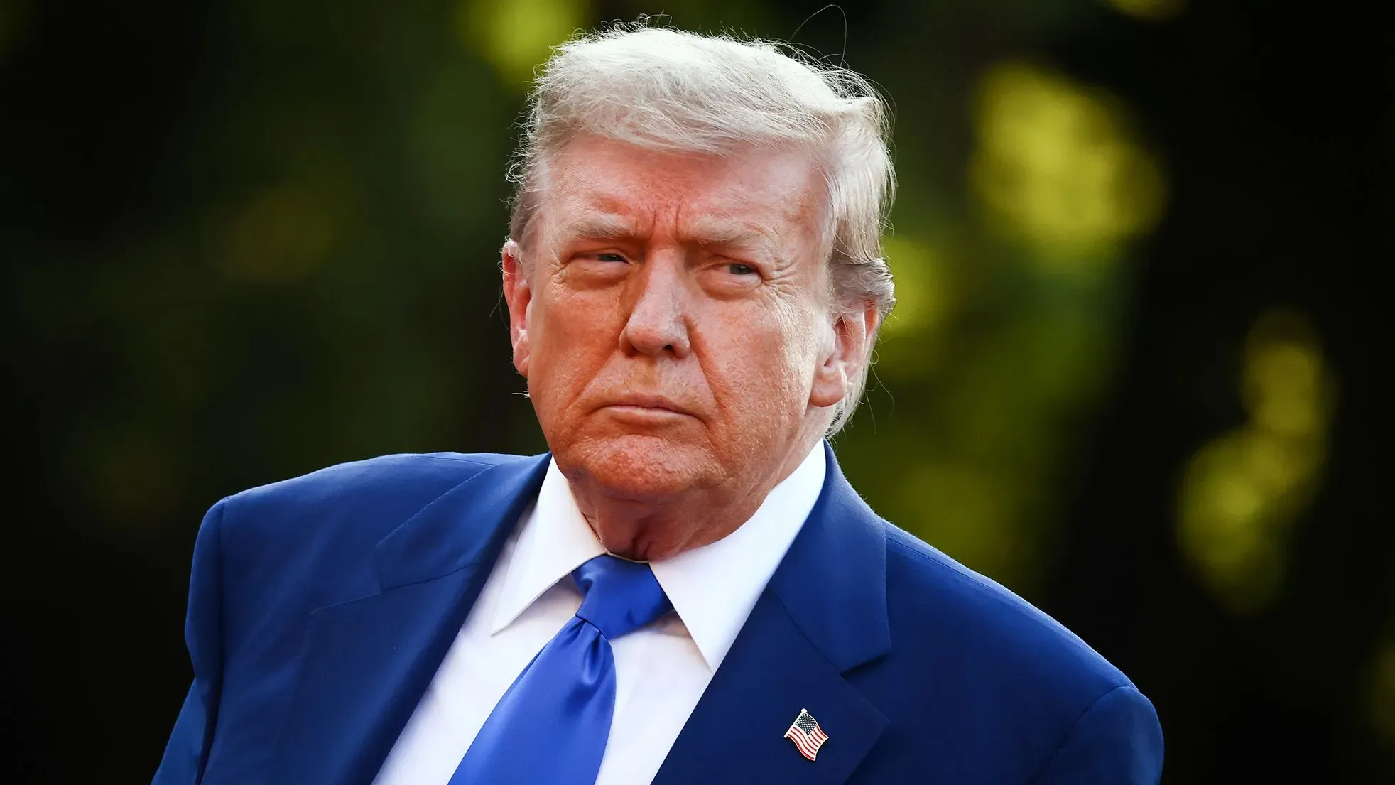 ترامب يظهر بملامح جدية، مرتديًا بدلة زرقاء، مع خلفية طبيعية، خلال قمة الناتو في هولندا، حيث تركز النقاشات على زيادة الإنفاق الدفاعي.