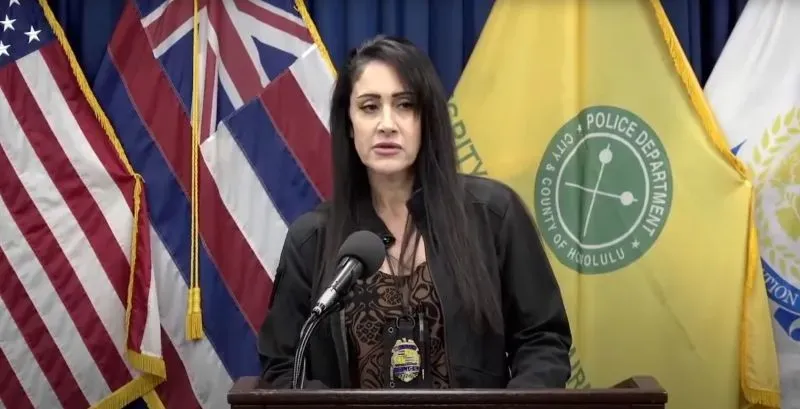 الملازم دينا ثويمز من شرطة هونولولو تتحدث في مؤتمر صحفي حول قضية قتل داون موموهارا، مع وجود أعلام هاواي والخدمات العامة خلفها.