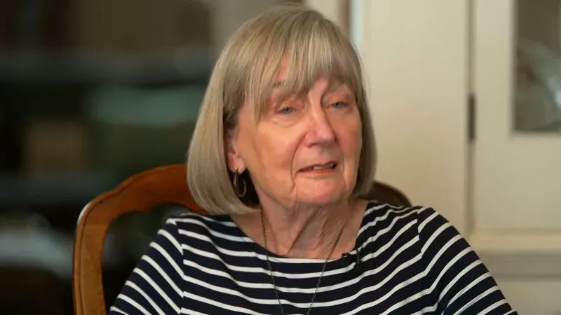 امرأة مسنّة ذات شعر رمادي قصير ترتدي قميصًا مخططًا بالأبيض والأسود، تعبر عن مشاعر القلق والارتباك أثناء حديثها عن تجربة زوجها مع إدارة الضمان الاجتماعي.