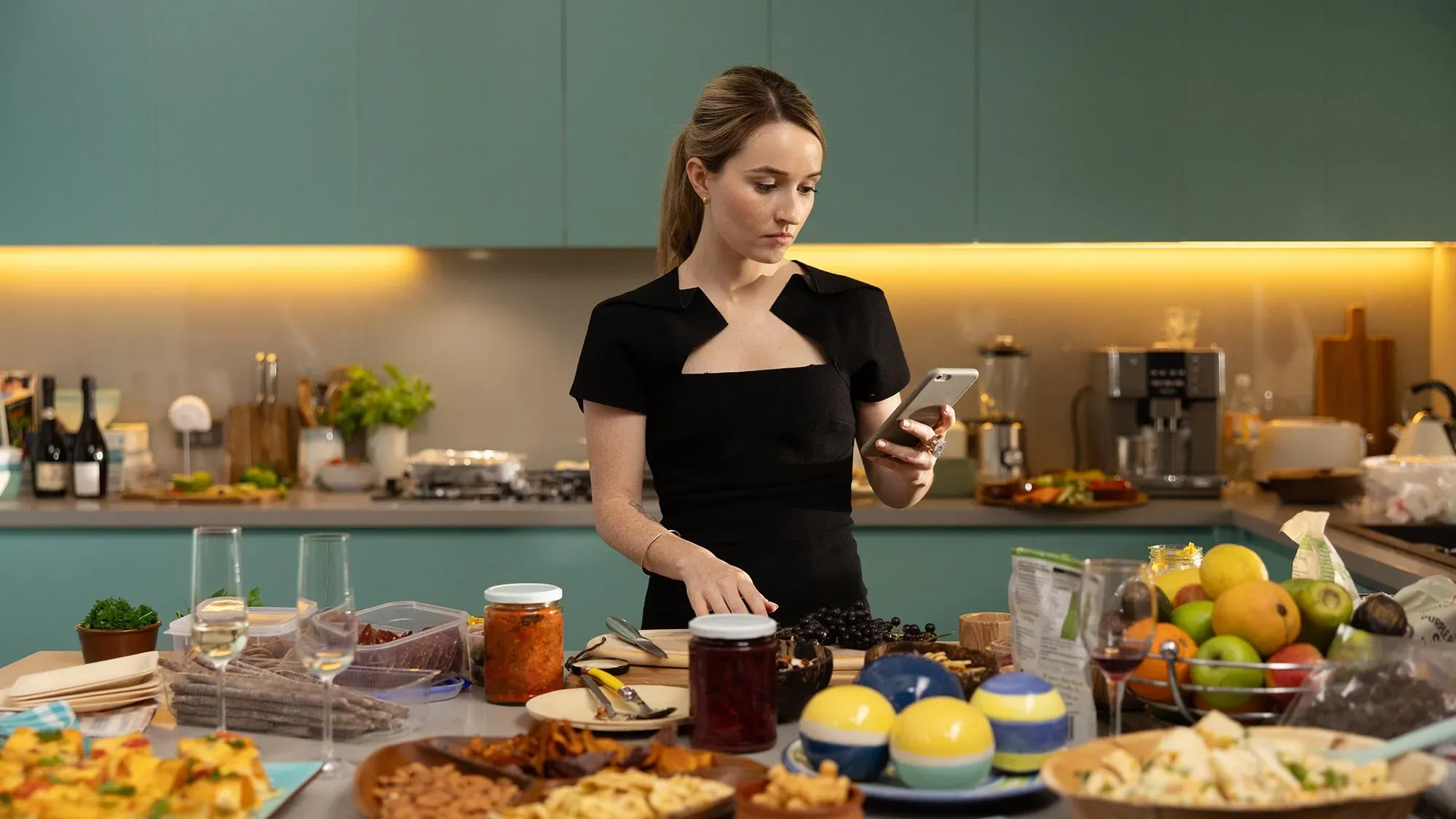 امرأة ترتدي فستانًا أسود، تقف في مطبخ عصري محاط بأطباق متنوعة من الطعام، بينما تتصفح هاتفها المحمول.