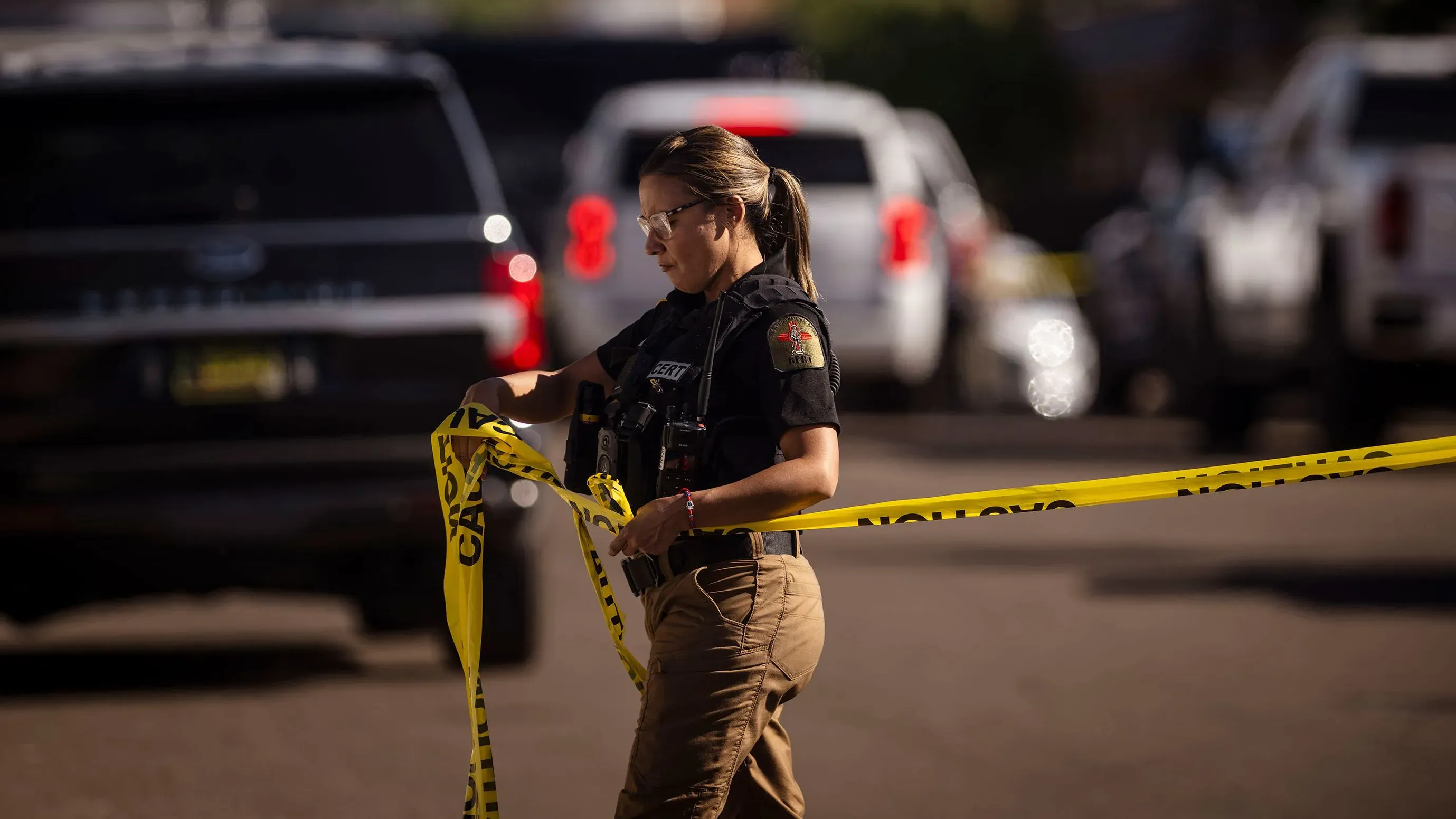 امرأة ترتدي زيًا رسميًا تحمل شريطًا تحذيريًا في موقع حادث، مع وجود سيارات الشرطة في الخلفية، تعكس جهود الحرس الوطني في تأمين المناطق.
