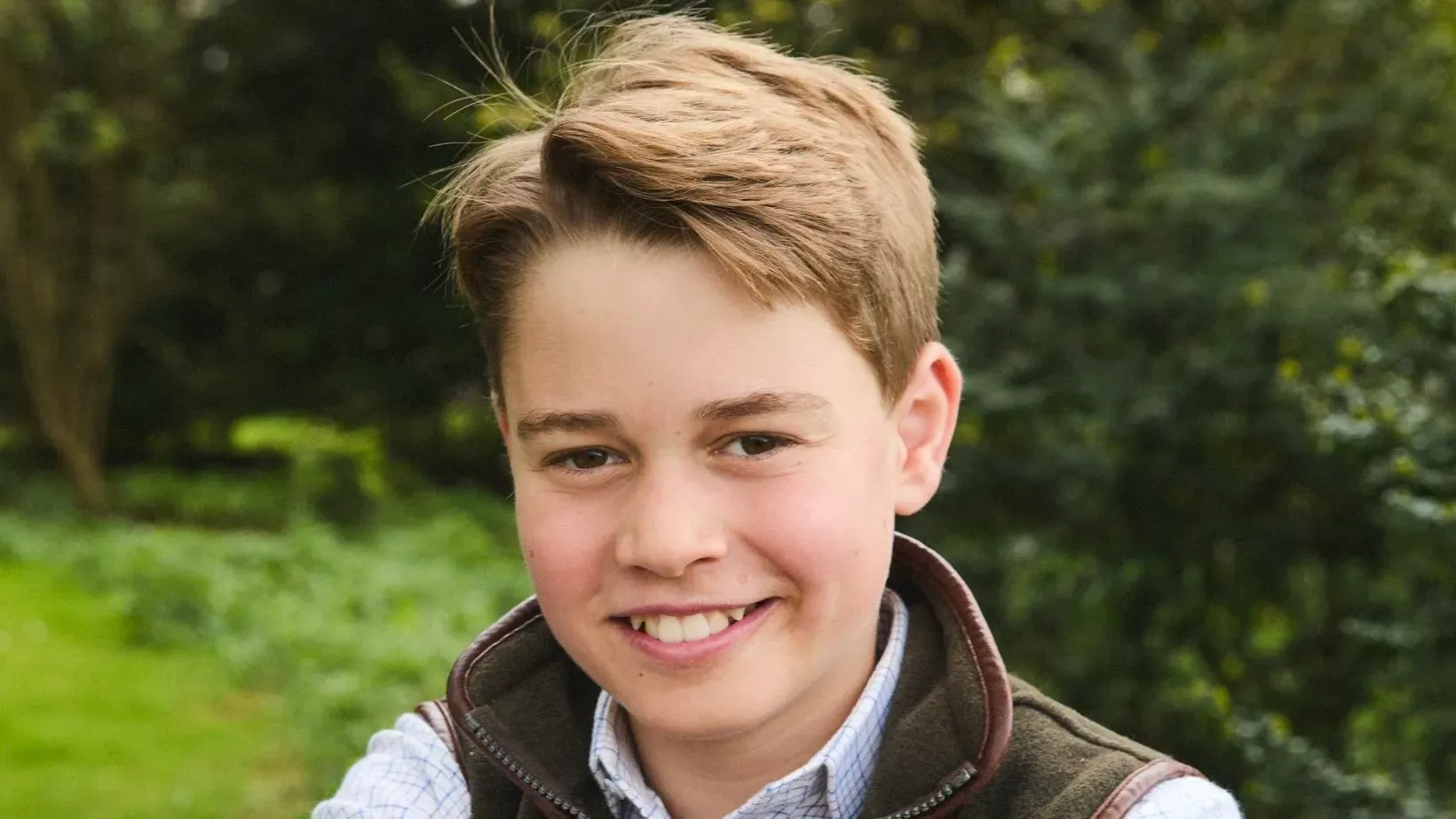 الأمير جورج، في عيد ميلاده الثاني عشر، مبتسم أمام بوابة خشبية في مكان ريفي، مرتديًا قميصًا أبيض وجيليه بني.