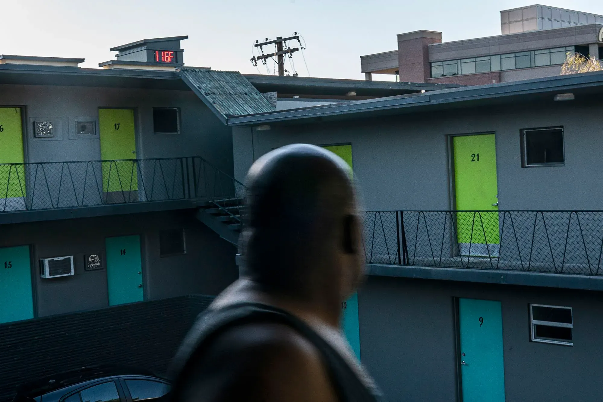 تظهر الصورة شخصًا يقف أمام فندق منخفض الارتفاع، مع أرقام الغرف وأبواب ملونة، مما يعكس تأثيرات تغير المناخ على المجتمعات.