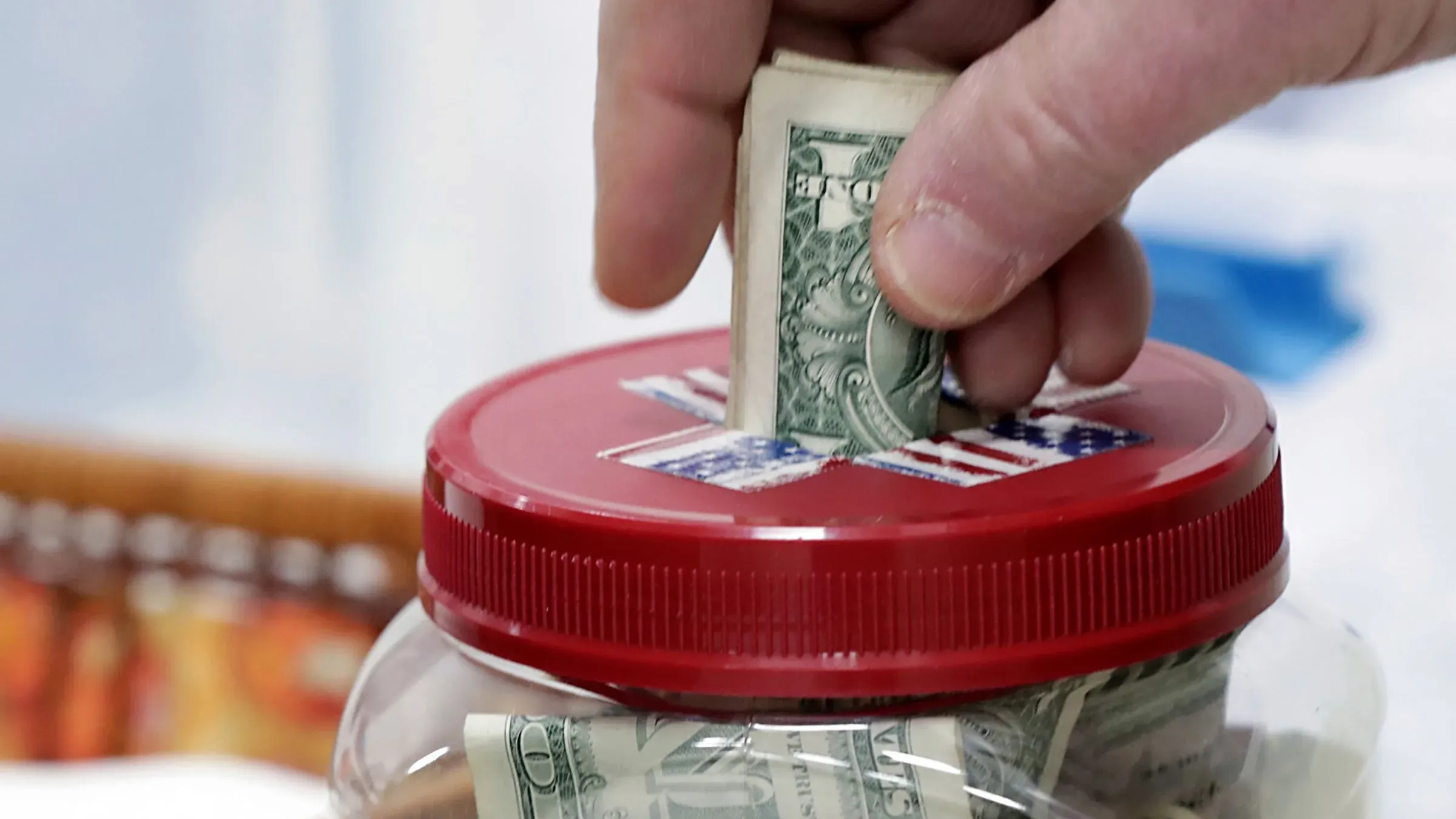يد تُدخل نقودًا في وعاء زجاجي لجمع التبرعات، مع وجود غطاء أحمر مزين بعلم أمريكي، مما يعكس أهمية التبرعات الخيرية.