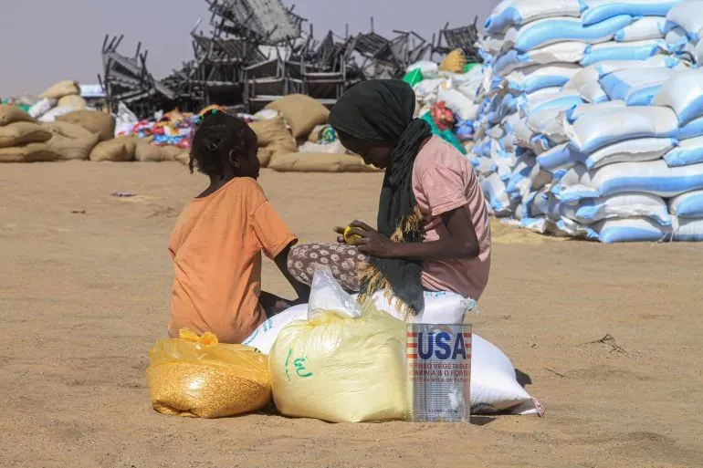 امرأتان جالستان على أكياس من المساعدات الغذائية في منطقة صحراوية، وسط أكياس أخرى، تعكسان أزمة إنسانية متزايدة في السودان.