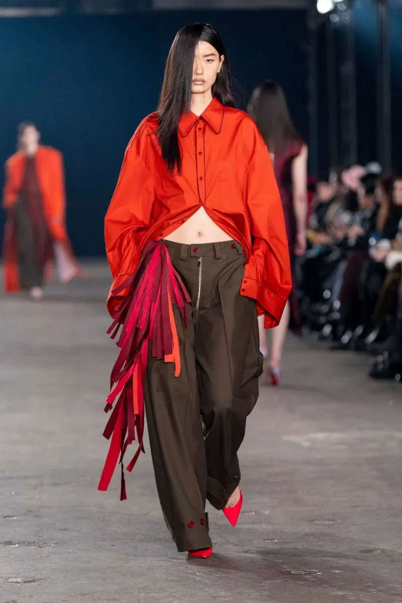 عارضة أزياء ترتدي قميصاً برتقالياً واسعاً وسروالاً بني اللون مزيناً بشرائط حمراء، تسير على ممشى عرض الأزياء في نيويورك.