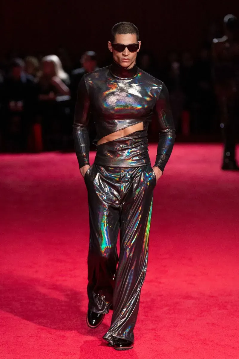 عارض أزياء يرتدي ملابس لامعة وعصرية، يسير على ممشى أحمر خلال عرض أزياء، مع نظارات شمسية تعكس أسلوب الموضة المعاصر.