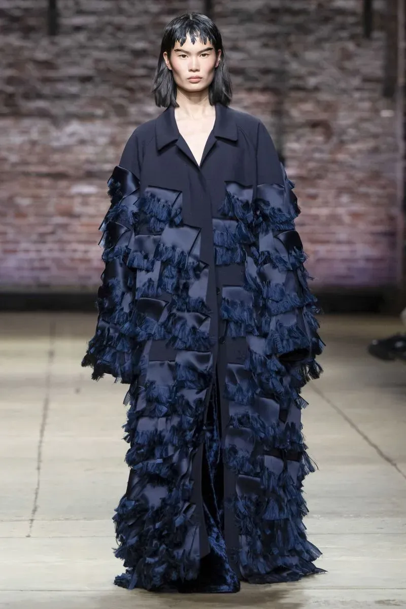 عرض أزياء يظهر عارضة ترتدي فستانًا بألوان داكنة مزخرف بالشرائط، في أسبوع الموضة بنيويورك، يعكس اتجاهات الموضة الحديثة.