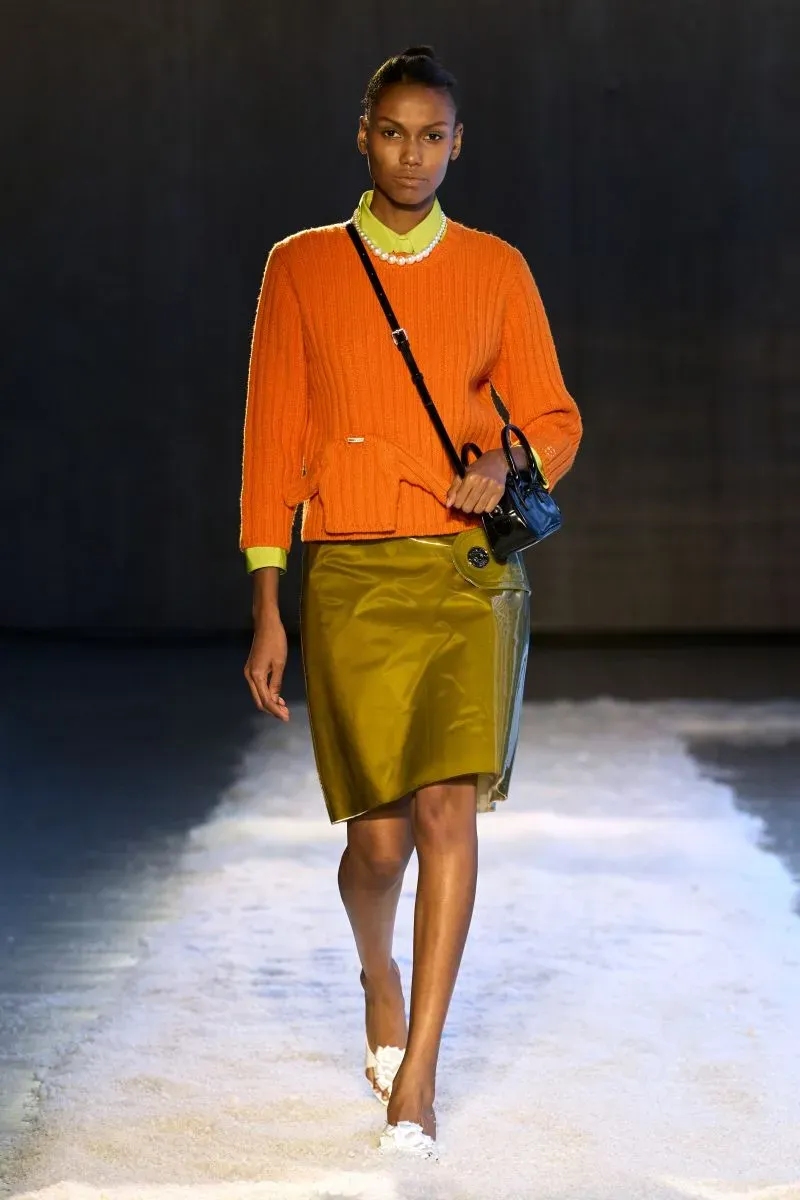 عارضة أزياء تسير على المنصة مرتدية سترة برتقالية وسكرتيرة صفراء، مع تنورة لامعة، في عرض أزياء حديث يعكس اتجاهات الموضة الأمريكية.