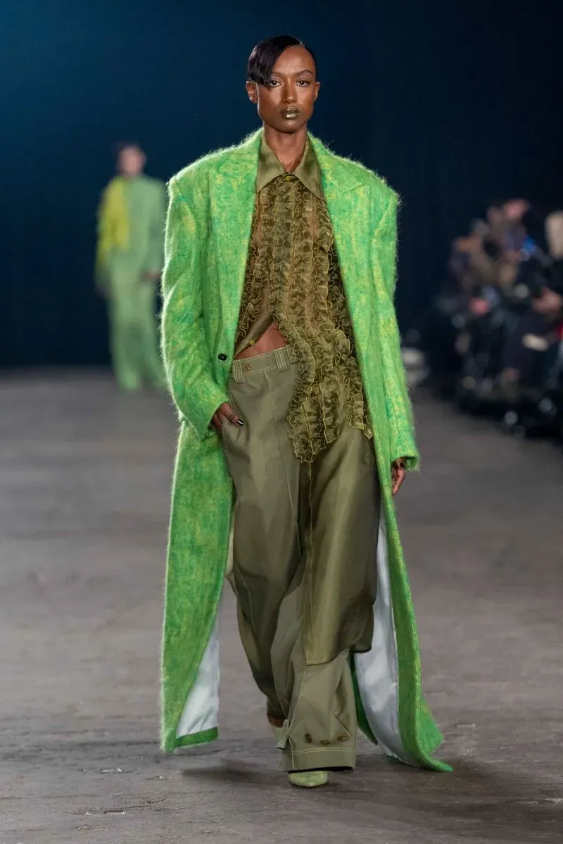 عارضة أزياء ترتدي معطفاً أخضر زاهياً مع تنسيق ألوان دافئة، تسير على منصة عرض أزياء في أسبوع الموضة بنيويورك، مع أجواء درامية.