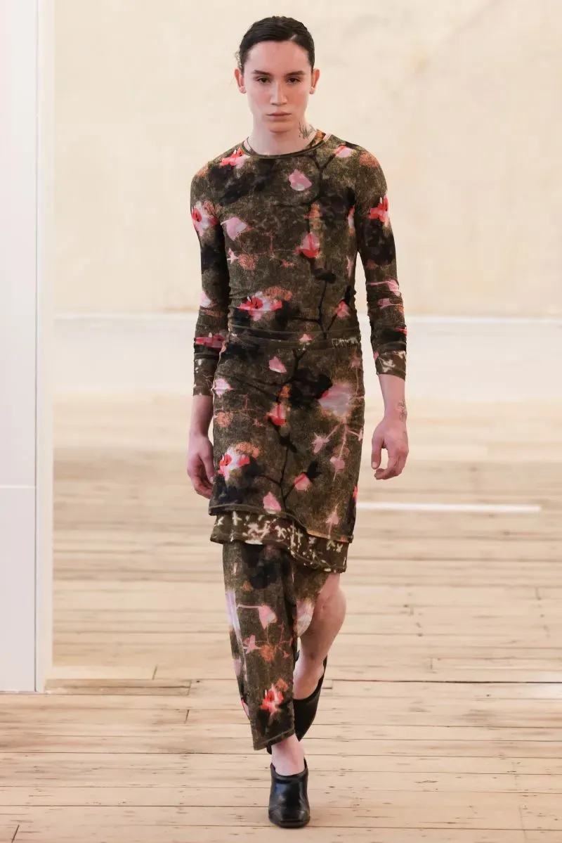 عارض أزياء يرتدي فستانًا مزخرفًا بألوان طبيعية، يمشي على منصة عرض خشبية، في إطار أسبوع الموضة بنيويورك.