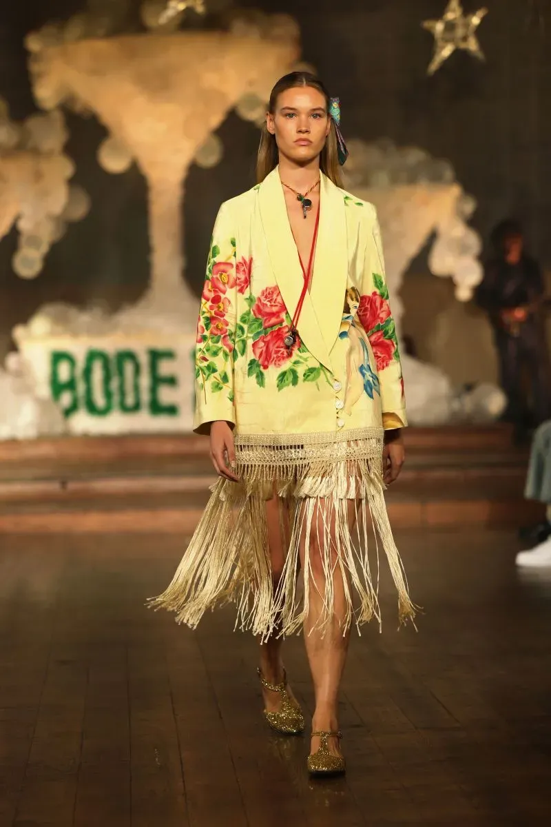 عارضة أزياء تسير على المنصة مرتدية جاكيت أصفر مزين بالورود مع تفاصيل من الخيوط، تعكس أحدث صيحات الموضة في أسبوع نيويورك.