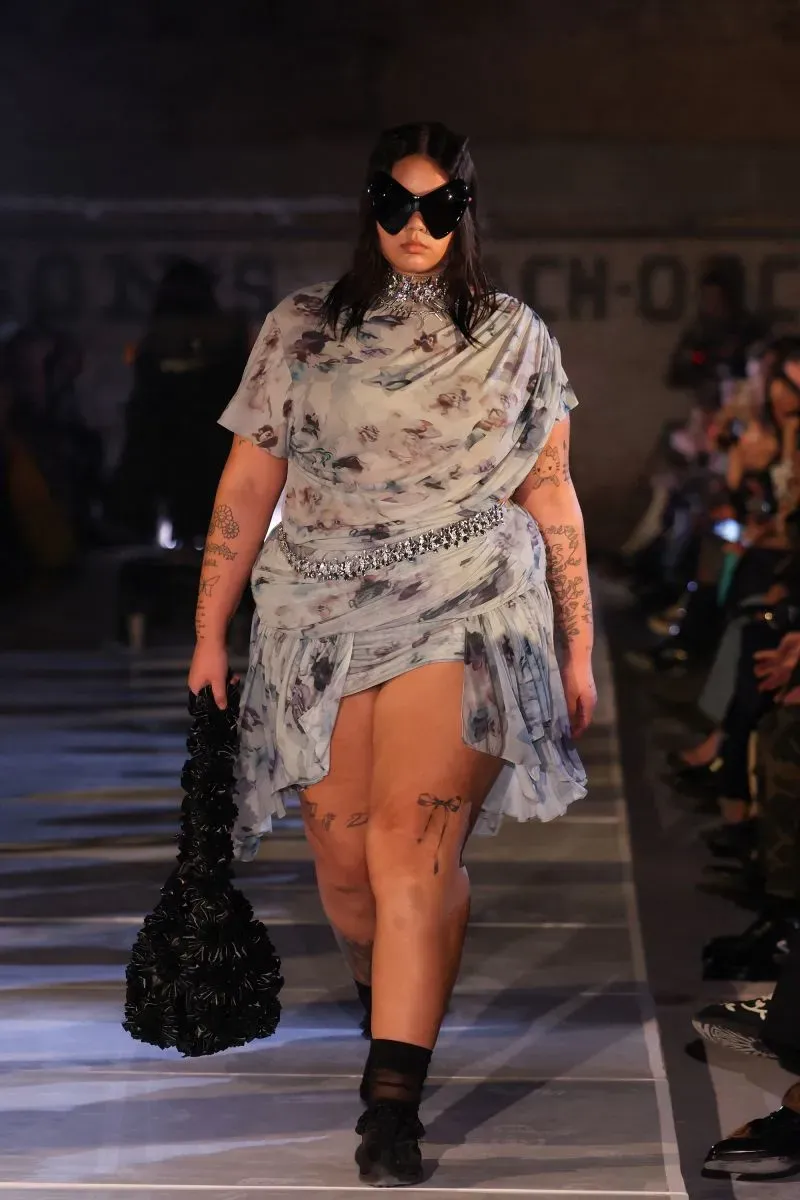 عارضة أزياء تسير على المدرج بفستان شفاف مزين برسومات، مع حقيبة سوداء كبيرة ونظارات شمسية، خلال أسبوع الموضة في نيويورك.