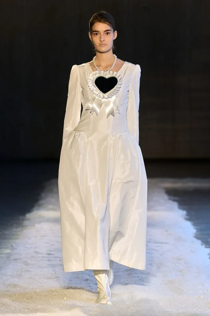 عارضة أزياء ترتدي فستانًا أبيض مزين بقلب أسود كبير، مع أكمام طويلة، تتجه نحو الكاميرا على منصة عرض الأزياء.