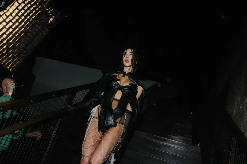 عارضة أزياء ترتدي فستاناً أسود جريئاً مع تفاصيل شبكية، تتجه نحو الكاميرا في أجواء مظلمة، مما يعكس اتجاهات الموضة المعاصرة.