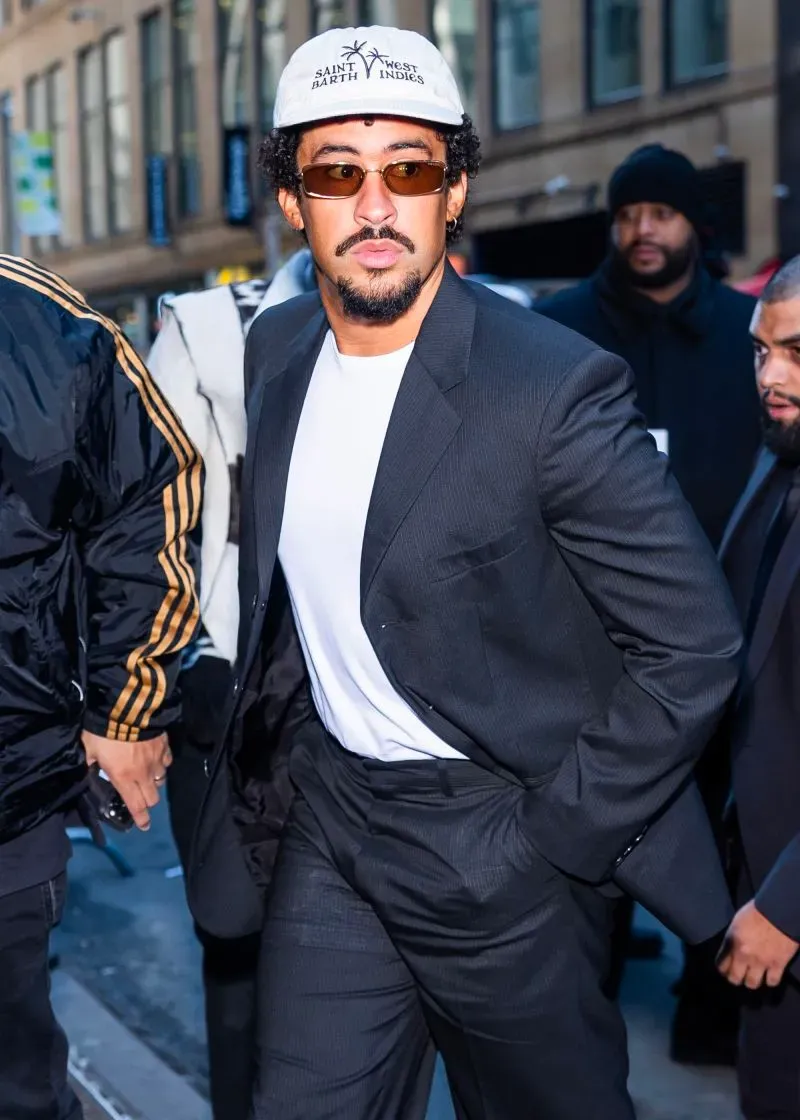 رجل يرتدي بدلة سوداء وقميص أبيض وقبعة، يسير في الشارع محاطًا بأشخاص آخرين، مع أجواء أسبوع الموضة في نيويورك.