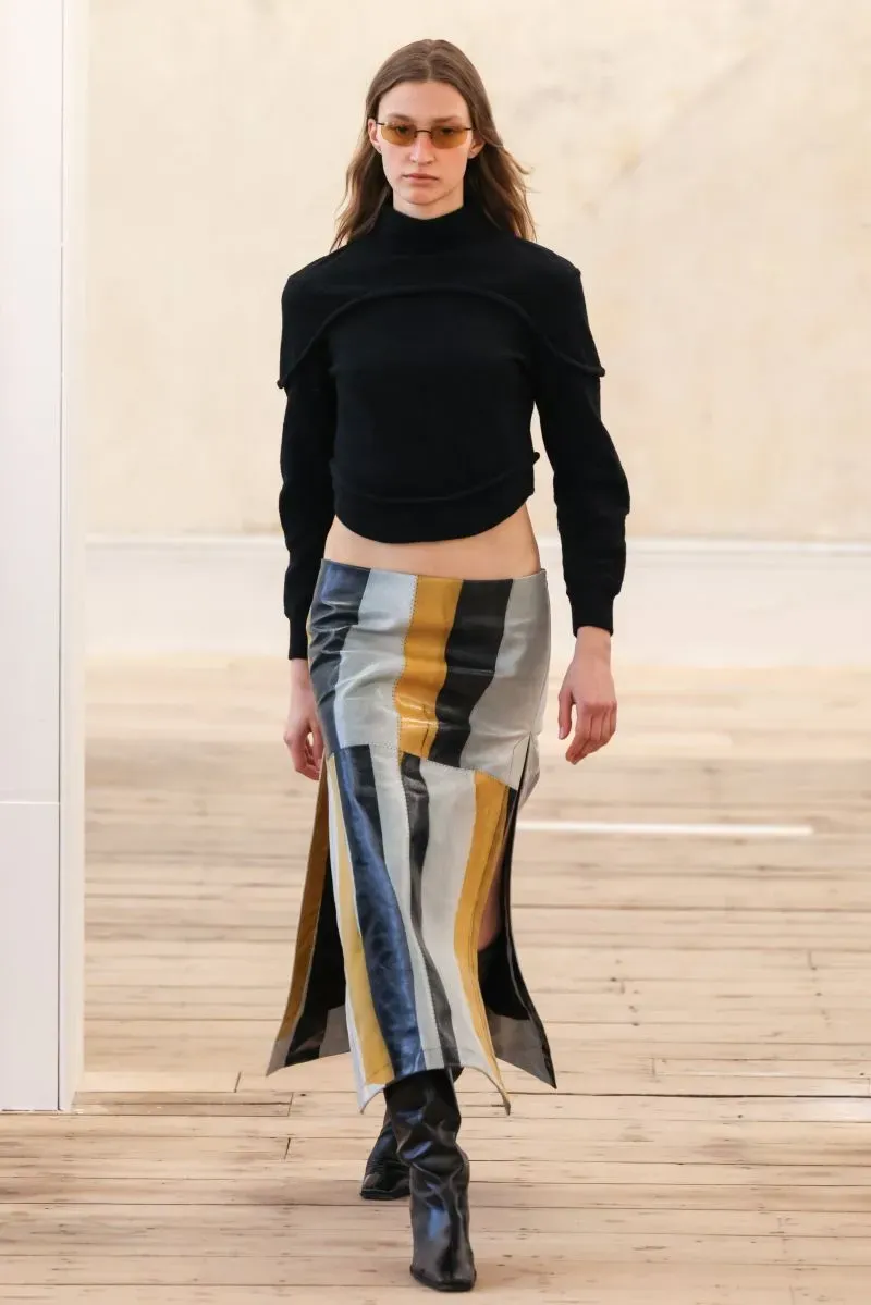 عارضة أزياء تسير على المنصة بفستان مزخرف بخطوط ملونة، مع سترة سوداء وقبعة شمسية، تعكس اتجاهات الموضة الحديثة في نيويورك.