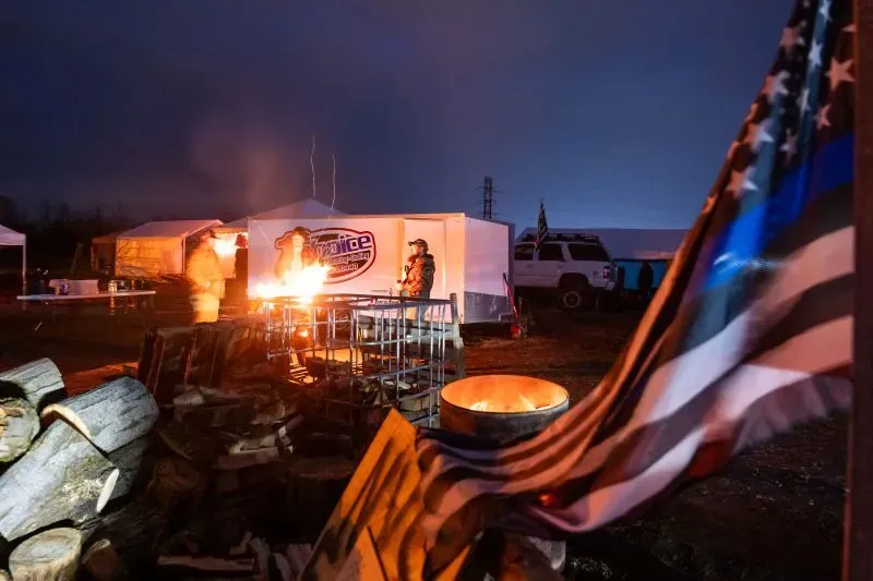 مشهد ليلي لمخيم إضراب ضباط الإصلاحيات في نيويورك، يظهر أشخاصًا حول نار، مع خيمة تحمل شعار الإضراب وعلم مميز.