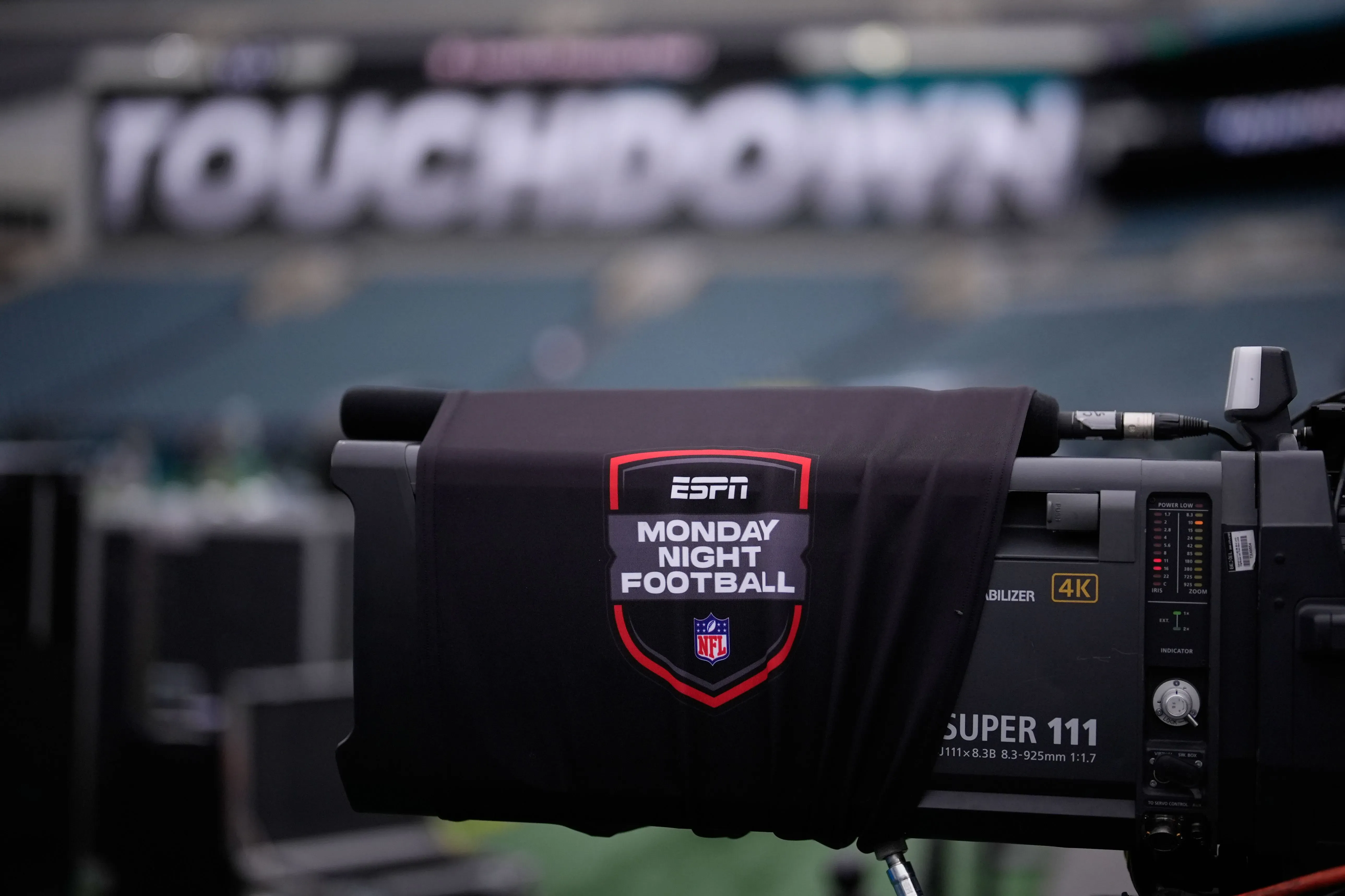 كاميرا تلفزيونية تحمل شعار "ESPN Monday Night Football" في ملعب كرة القدم، مع خلفية تظهر كلمة "TOUCHDOWN".