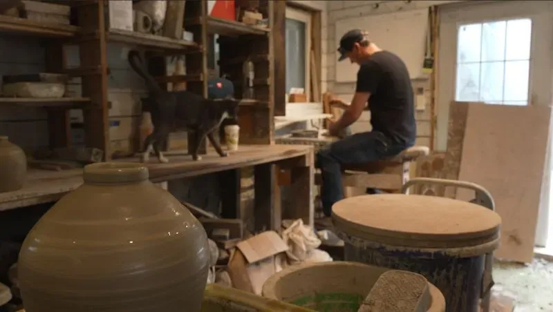 رجل يعمل في ورشة فخارية، بينما يتواجد قطة سوداء بجانبه، مع أدوات فخارية وأوانٍ حوله. تعكس الصورة أجواء الإبداع والحرفية.