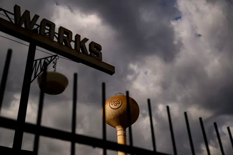 صورة لمصنع يو إس ستيل تحت سماء غائمة، مع وجود خزان مياه يحمل شعار الشركة، مما يعكس تحديات الصناعة الأمريكية.