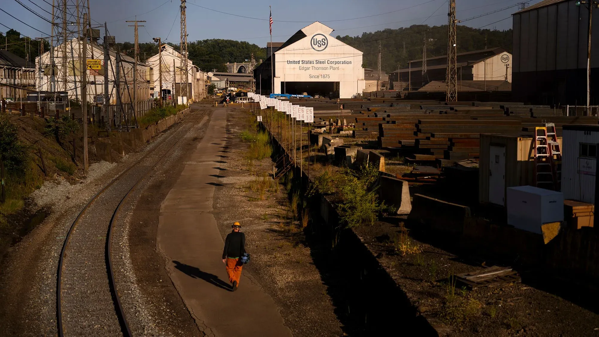 مشهد لمصنع يو إس ستيل، يظهر شخصًا يسير في ممر بجوار المباني الصناعية، مع لافتة تحمل اسم الشركة، مما يعكس التحديات التي تواجهها الشركة.