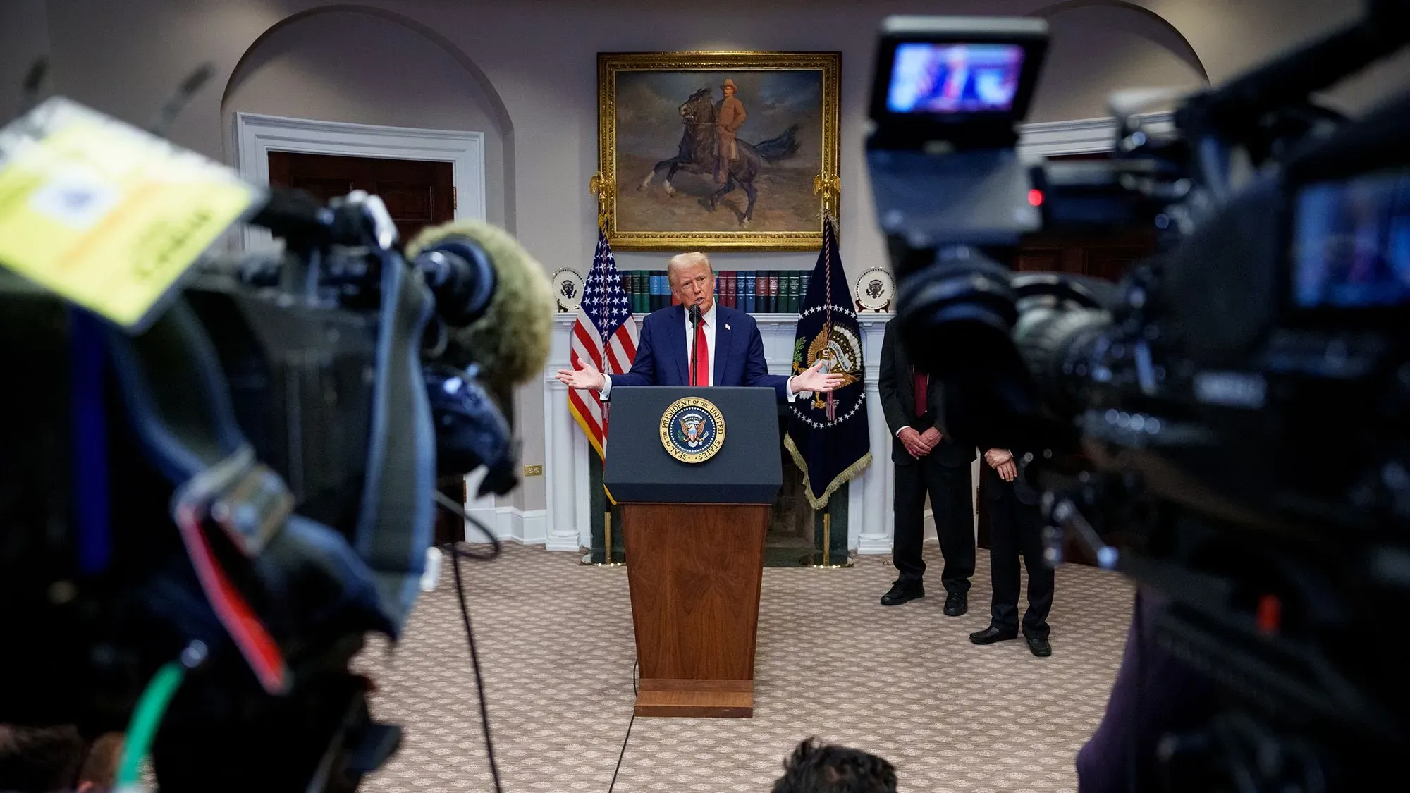 ترامب يتحدث في مؤتمر صحفي بالبيت الأبيض، مع كاميرات الإعلام والأعلام الأمريكية خلفه، وسط خلفية فنية تاريخية.