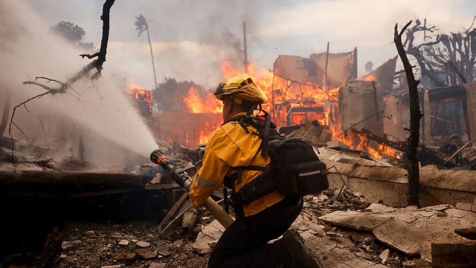 رجل إطفاء يستخدم خرطومًا لمكافحة حرائق الغابات في كاليفورنيا، بينما تتصاعد ألسنة اللهب من المباني المدمرة خلفه.
